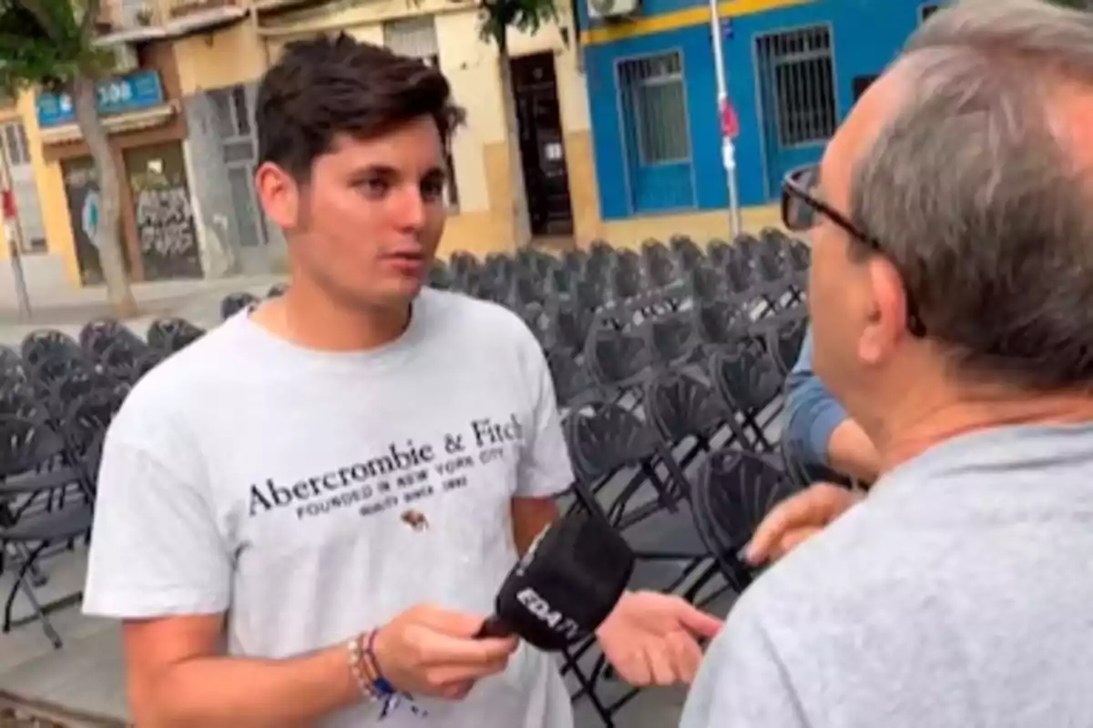 Un joven con una camiseta de Abercrombie & Fitch sostiene un micrófono mientras conversa con un hombre mayor en una calle con sillas vacías al fondo.