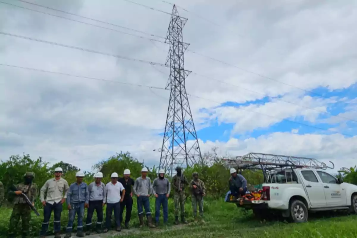 Un grupo de personas con cascos de seguridad y uniformes está de pie junto a un vehículo blanco en un área verde con una torre de electricidad al fondo.