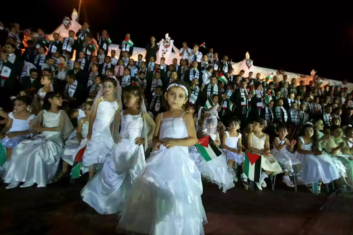 Un grupo de niñas vestidas de blanco y niños con trajes formales sostienen banderas en un evento nocturno.