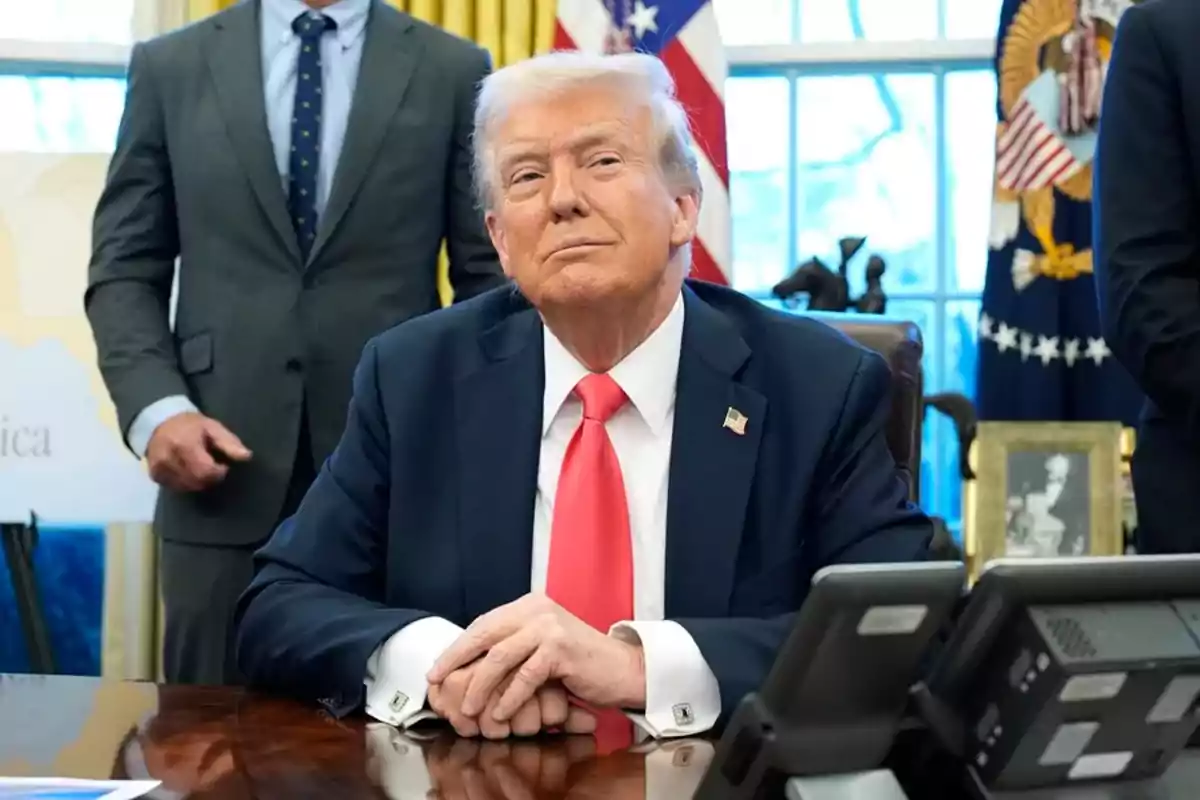 A man in a dark suit and red tie is sitting at a desk with his hands clasped, while two people stand behind him in an office with flags and an official seal on the wall.