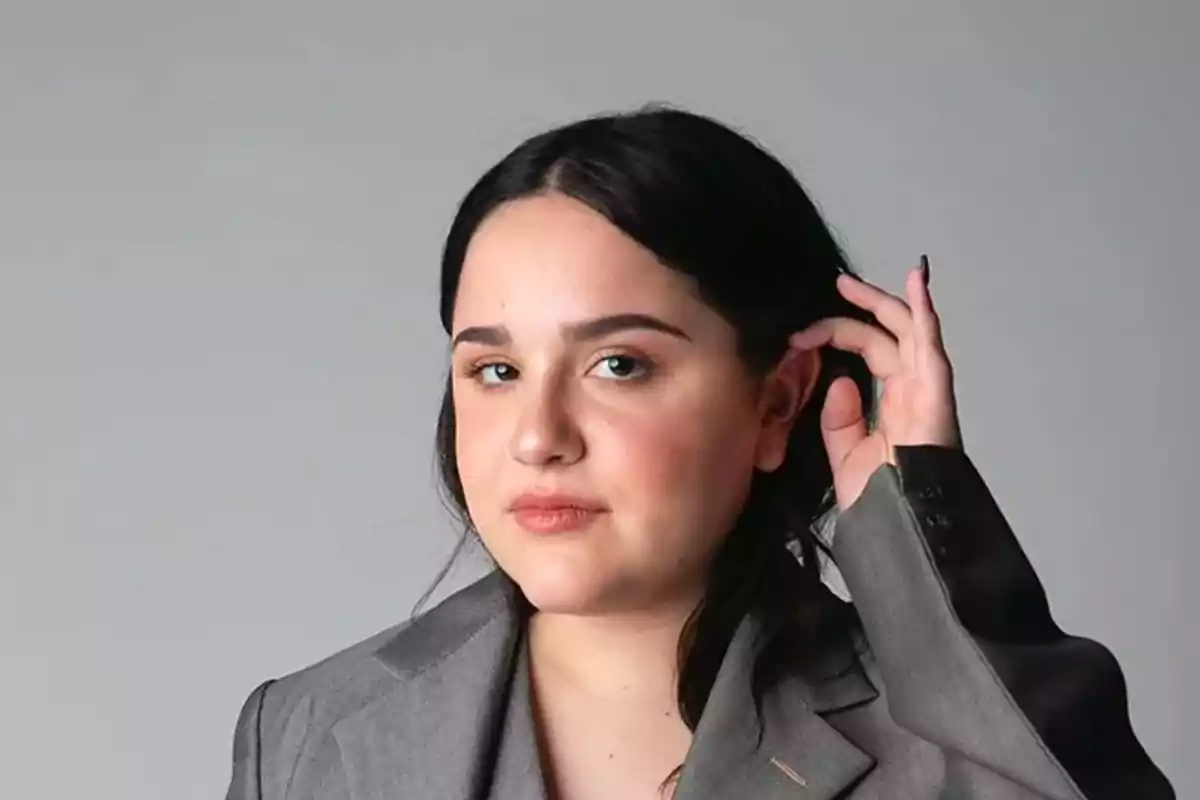 Mujer con cabello oscuro y traje gris tocándose el cabello frente a un fondo gris.