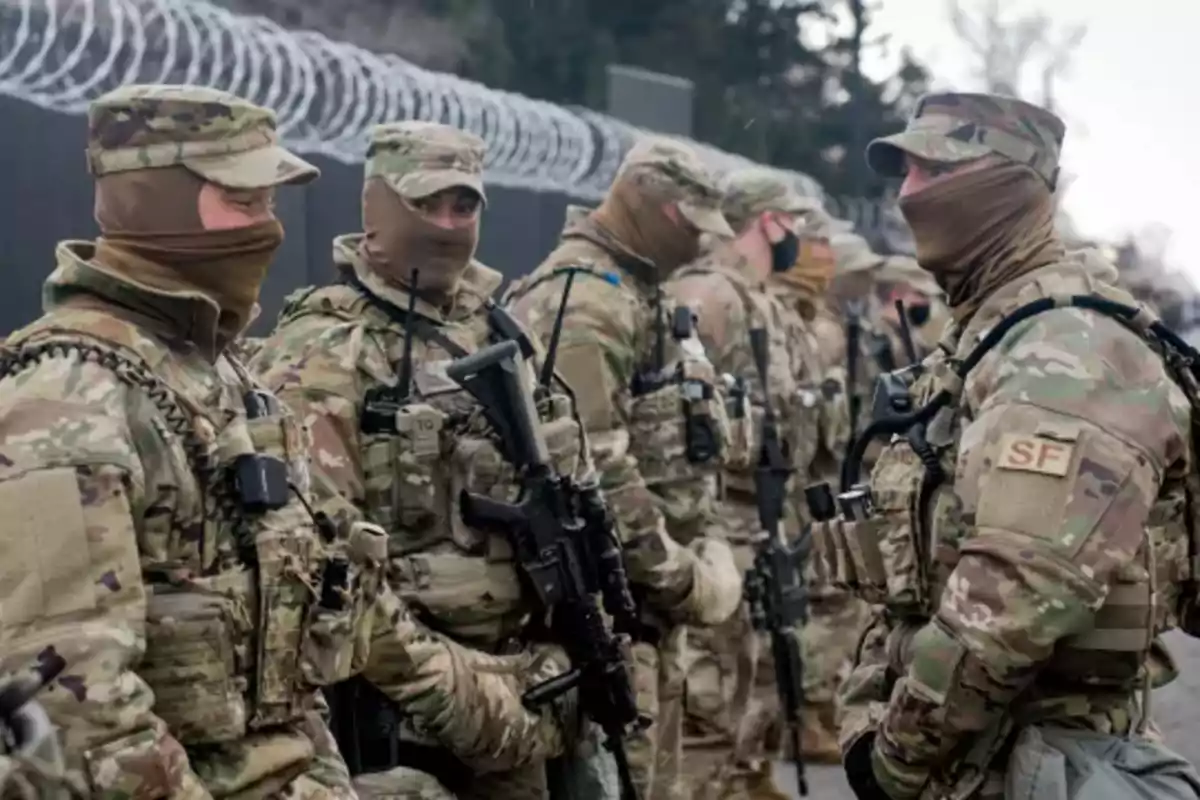 Soldados con uniforme de camuflaje y pasamontañas en formación junto a una cerca de alambre de púas.