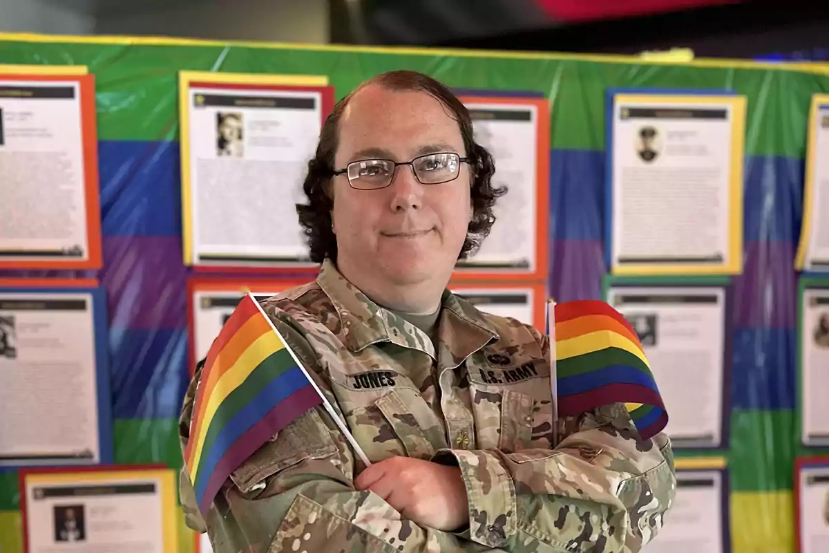 Una persona en uniforme militar sostiene dos banderas del orgullo LGBTQ+ frente a un fondo colorido con documentos enmarcados.