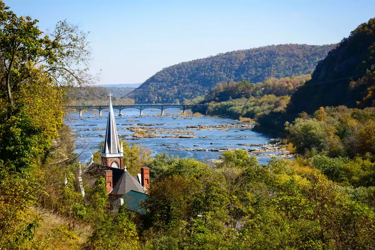 Una iglesia con un campanario se encuentra rodeada de árboles con un río y un puente al fondo, en un paisaje montañoso.