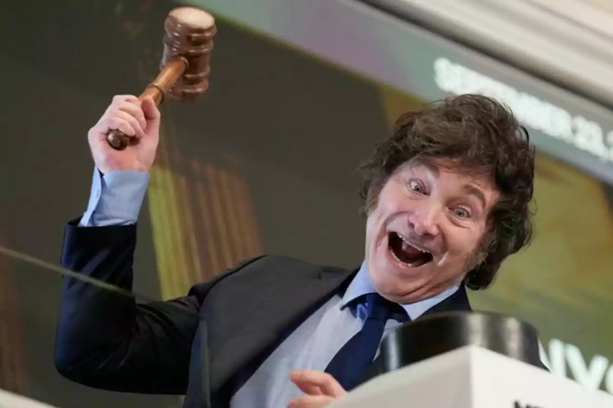 Un hombre sonriente con un mazo en la mano, vestido con traje y corbata, parece estar en un evento o ceremonia.