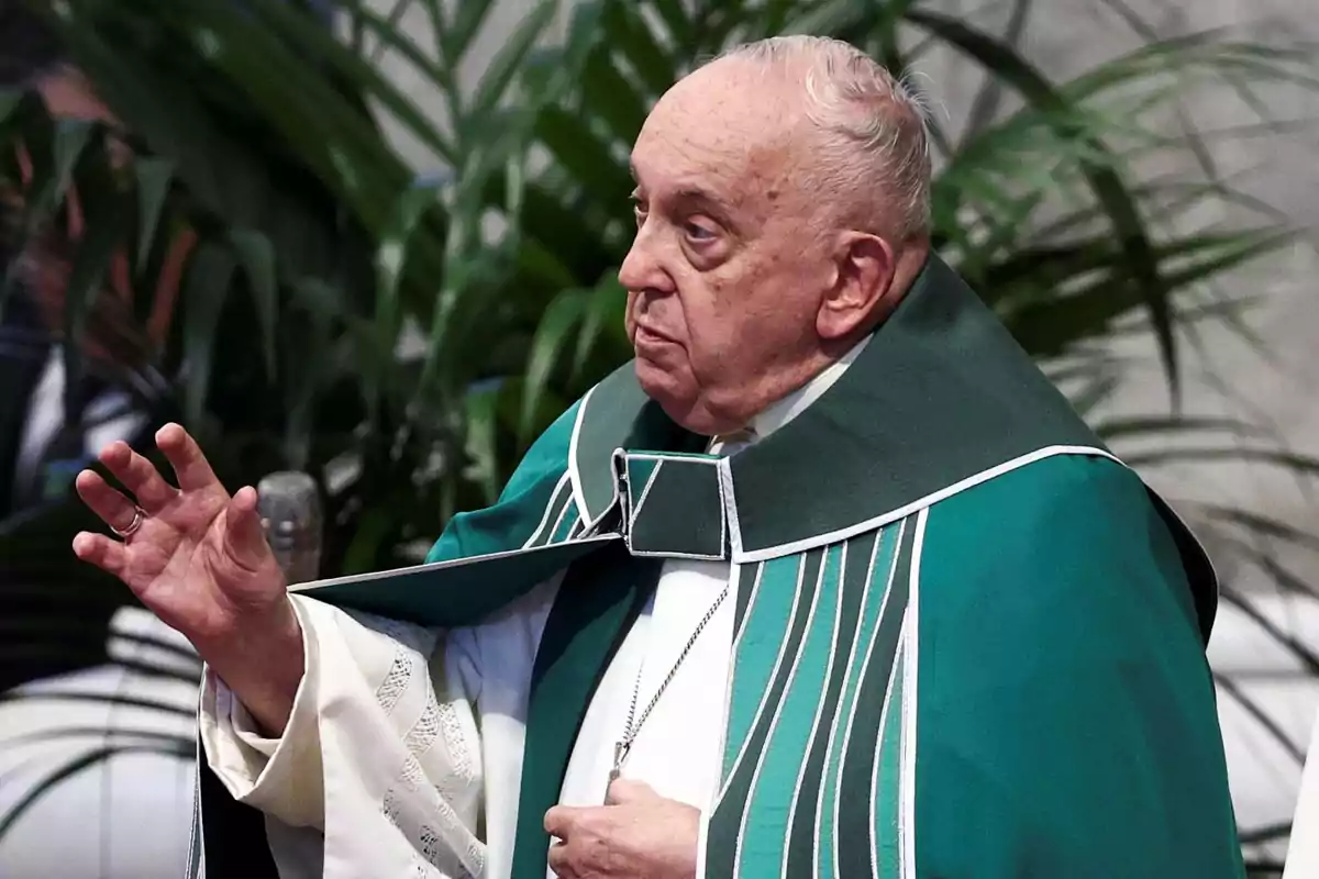 An elderly man dressed in a green and white robe, raising one hand, with plants in the background.