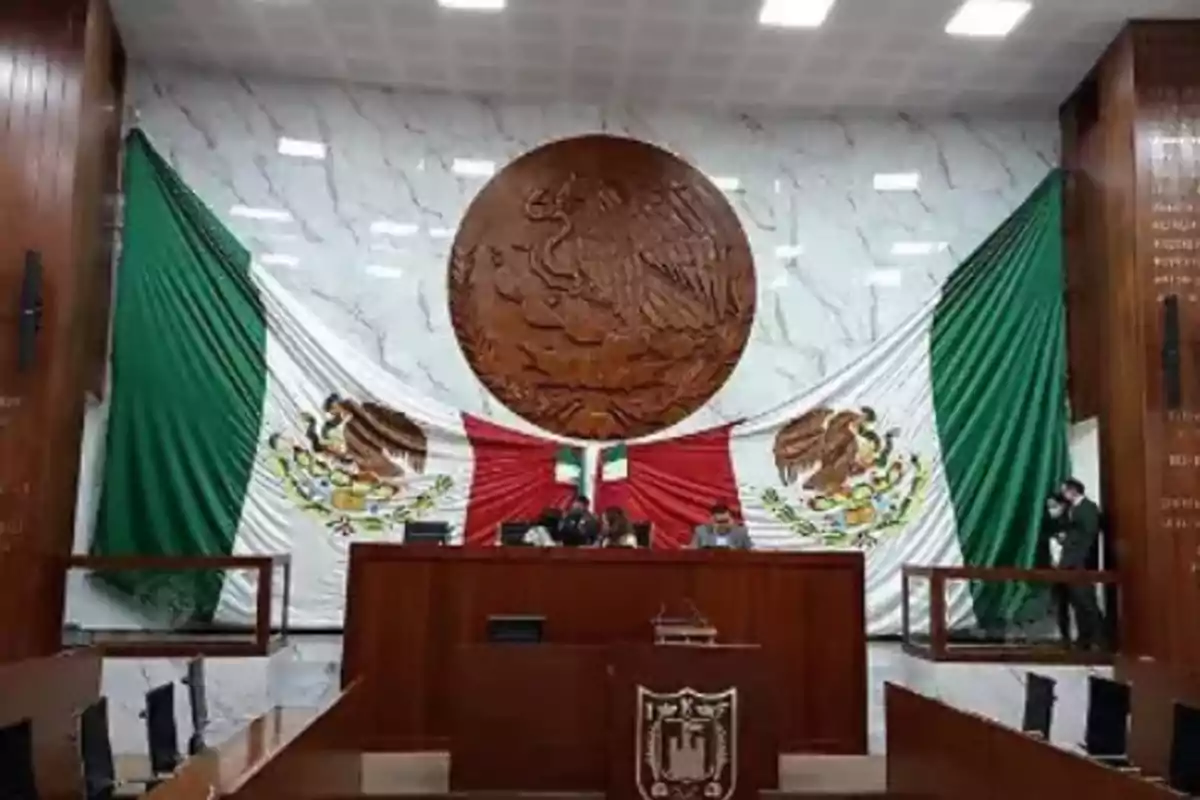 Session hall with a large Mexican flag and a coat of arms on the wall behind a podium.