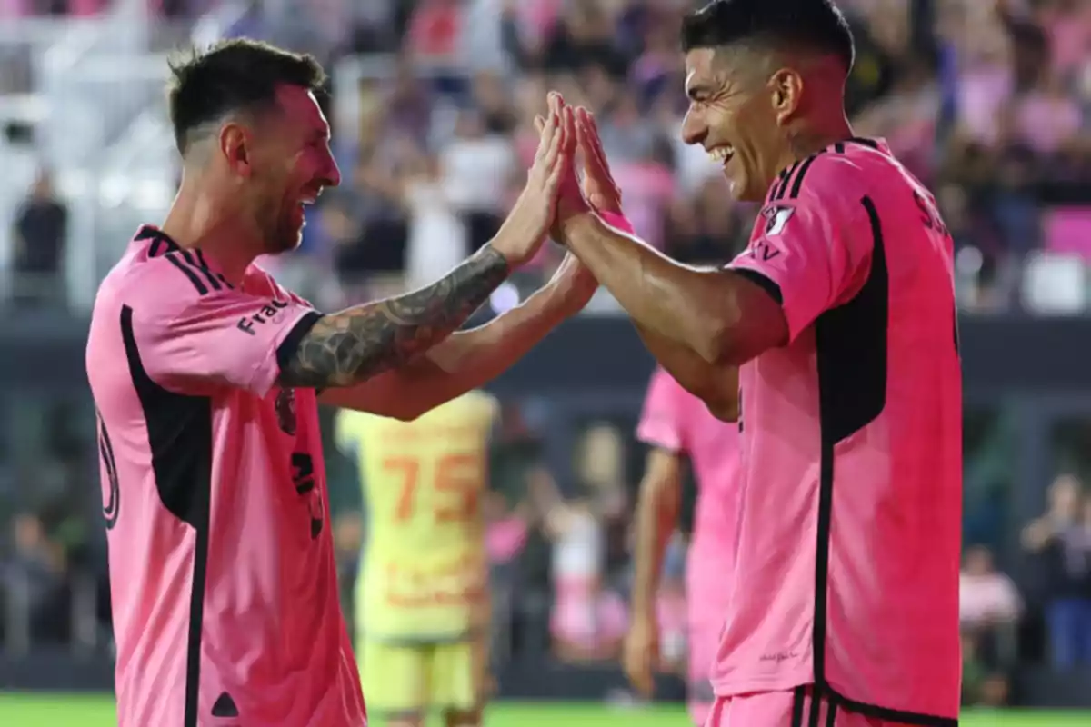 Dos jugadores de fútbol con uniformes rosas celebran en el campo con una sonrisa y un choque de manos.