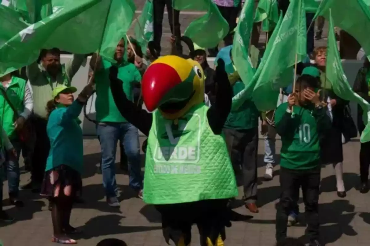 Un grupo de personas vestidas de verde ondea banderas mientras una mascota con forma de tucán se encuentra en el centro.