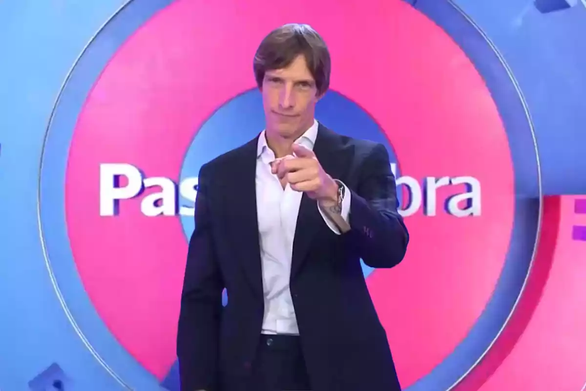 Ivan de Pineda de traje oscuro y camisa blanca señala hacia adelante con un fondo colorido que tiene texto parcialmente visible de "Pasapalabra".