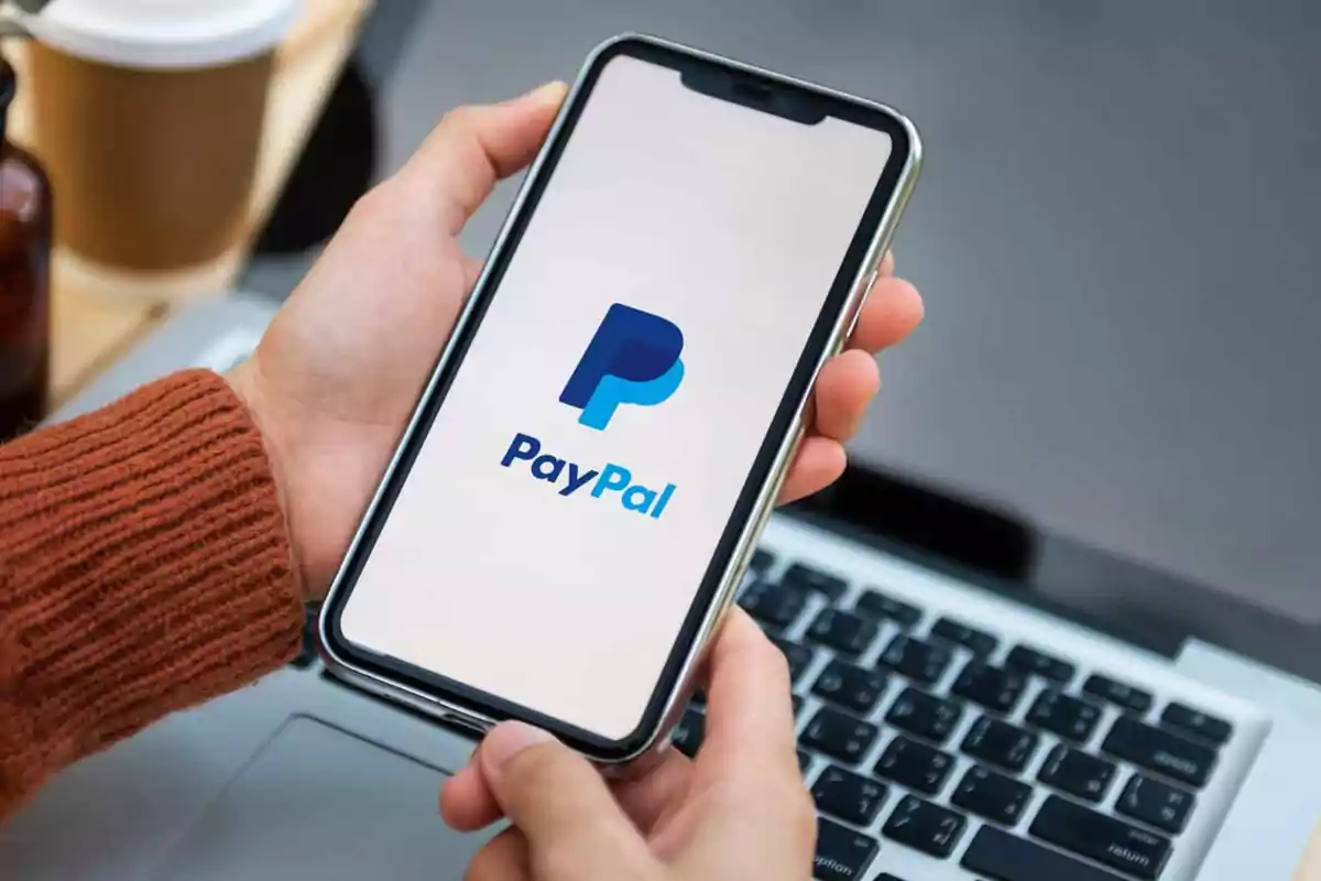 Persona sosteniendo un teléfono móvil con el logo de PayPal en la pantalla frente a una computadora portátil.