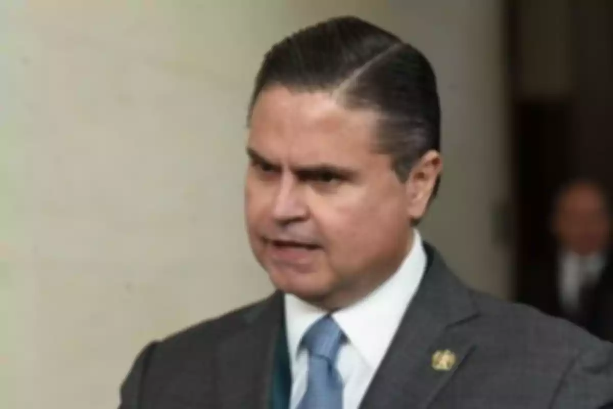 A man in a dark suit and blue tie speaking in a formal setting.