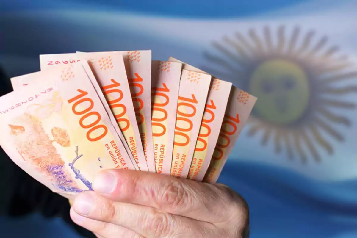 Una mano sosteniendo billetes de mil pesos argentinos con la bandera de Argentina de fondo.