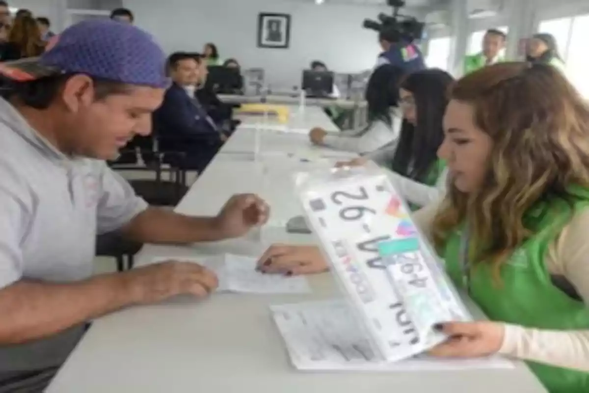 A man and a woman are sitting in an office, the woman hands the man a package that seems to contain license plates.