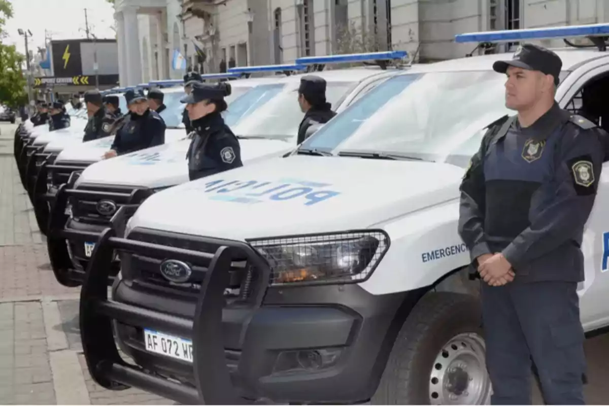 Policías uniformados de pie junto a una fila de vehículos policiales Ford estacionados en una calle.