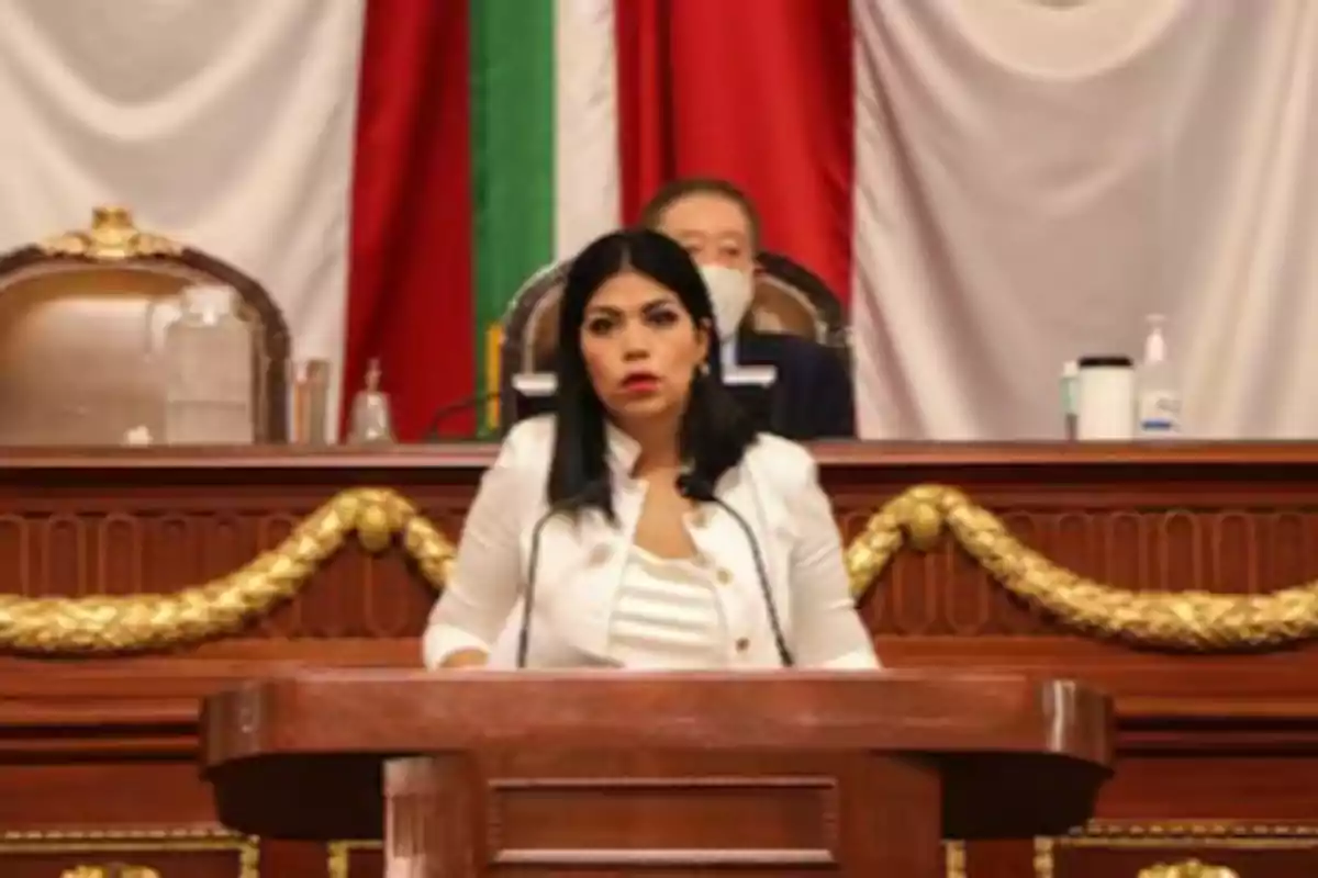 A woman standing at a podium in a formal setting with flags and decorations behind her.