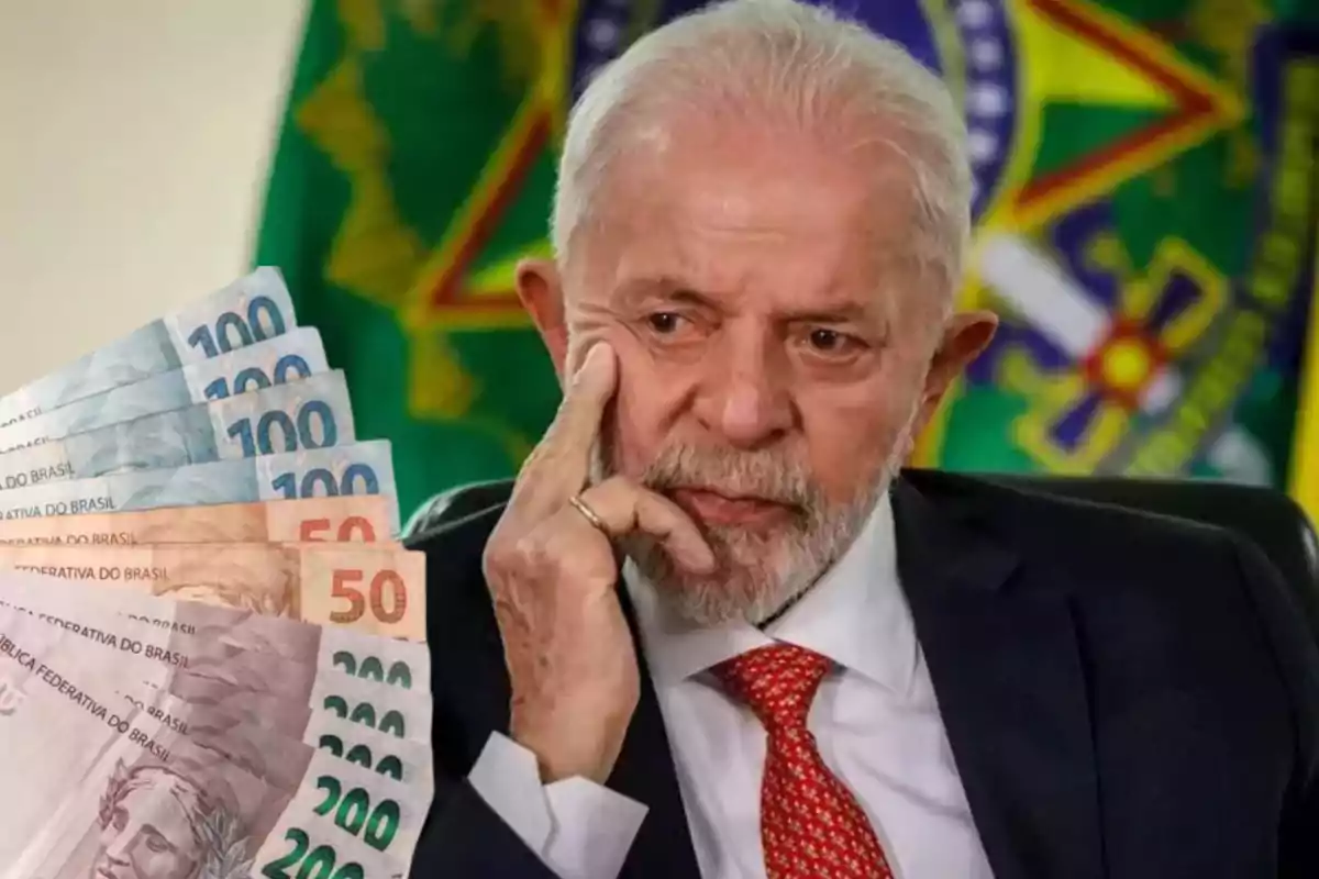 Un hombre mayor con traje y corbata roja está pensativo frente a un fondo con una bandera, mientras sostiene varios billetes de real brasileño.