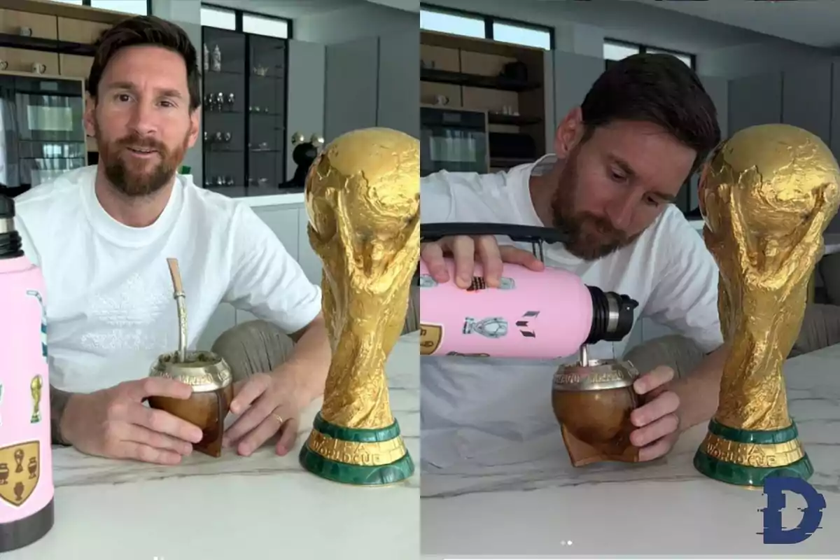 Un hombre con barba y camiseta blanca está sentado en una mesa con un trofeo dorado y un mate, mientras vierte agua caliente de un termo rosa en el mate.