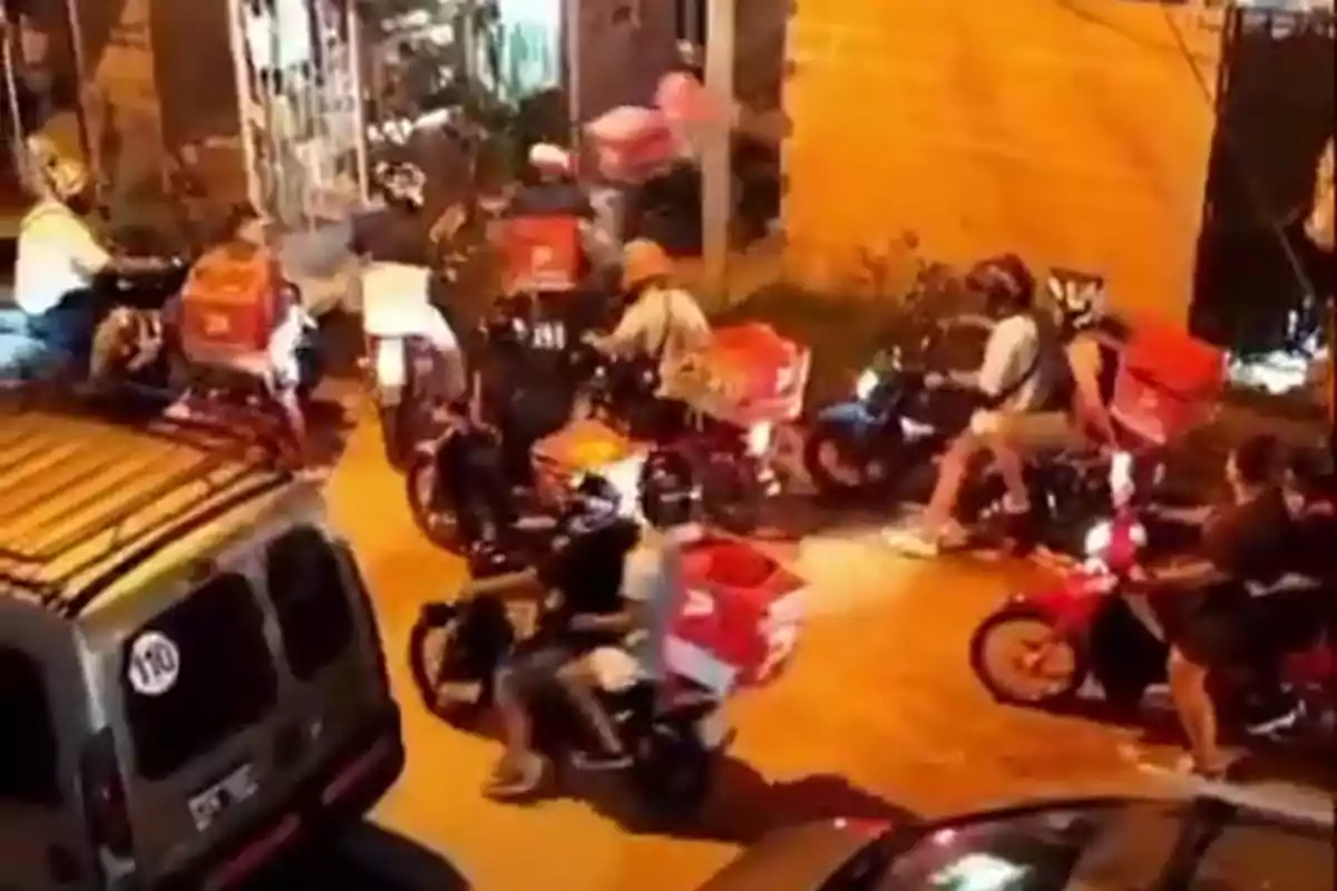 A group of motorcyclists with delivery boxes gathers on a street lit up at night.