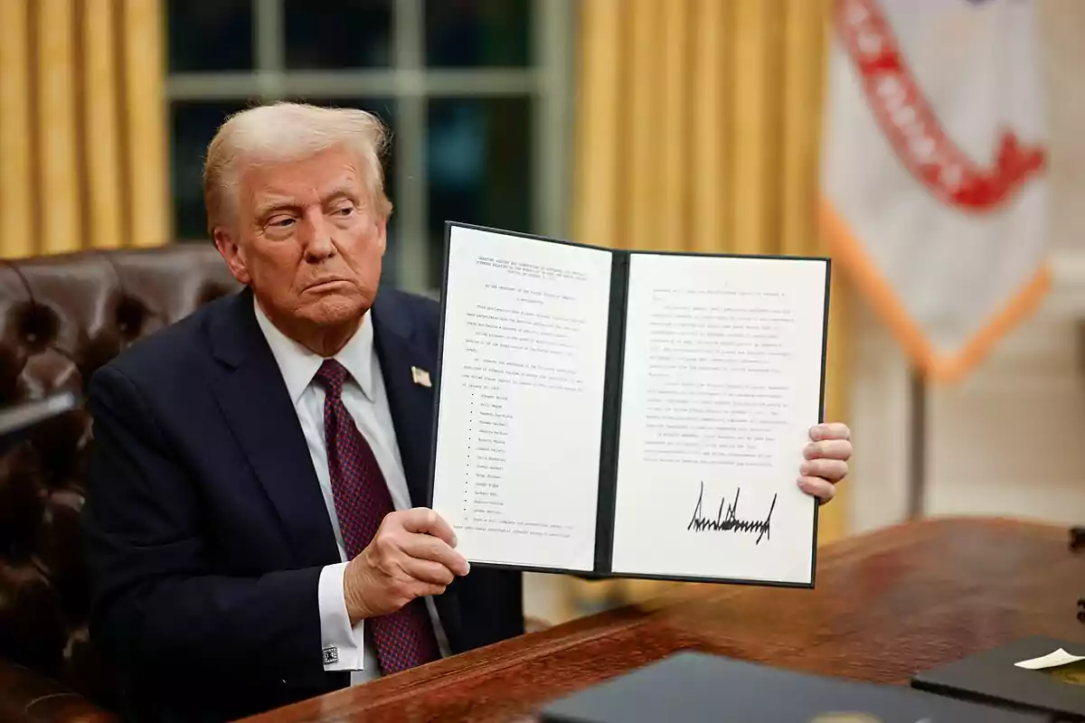 Un hombre de traje oscuro y corbata roja está sentado en un escritorio sosteniendo un documento abierto con texto y una firma visible.