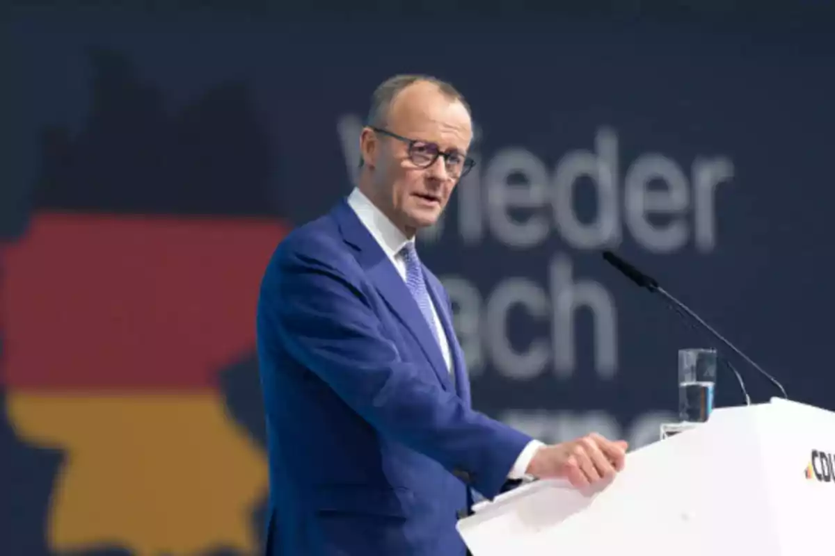 Un hombre con traje azul y gafas habla en un podio con un fondo que muestra un mapa de Alemania.