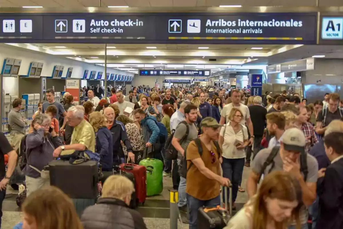 Una multitud de personas con maletas se encuentra en un aeropuerto, bajo señales que indican "Auto check-in" y "Arribos internacionales".