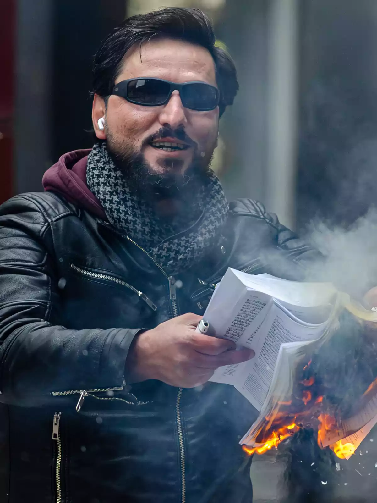 Un hombre con gafas de sol y auriculares sostiene un libro en llamas mientras sonríe.