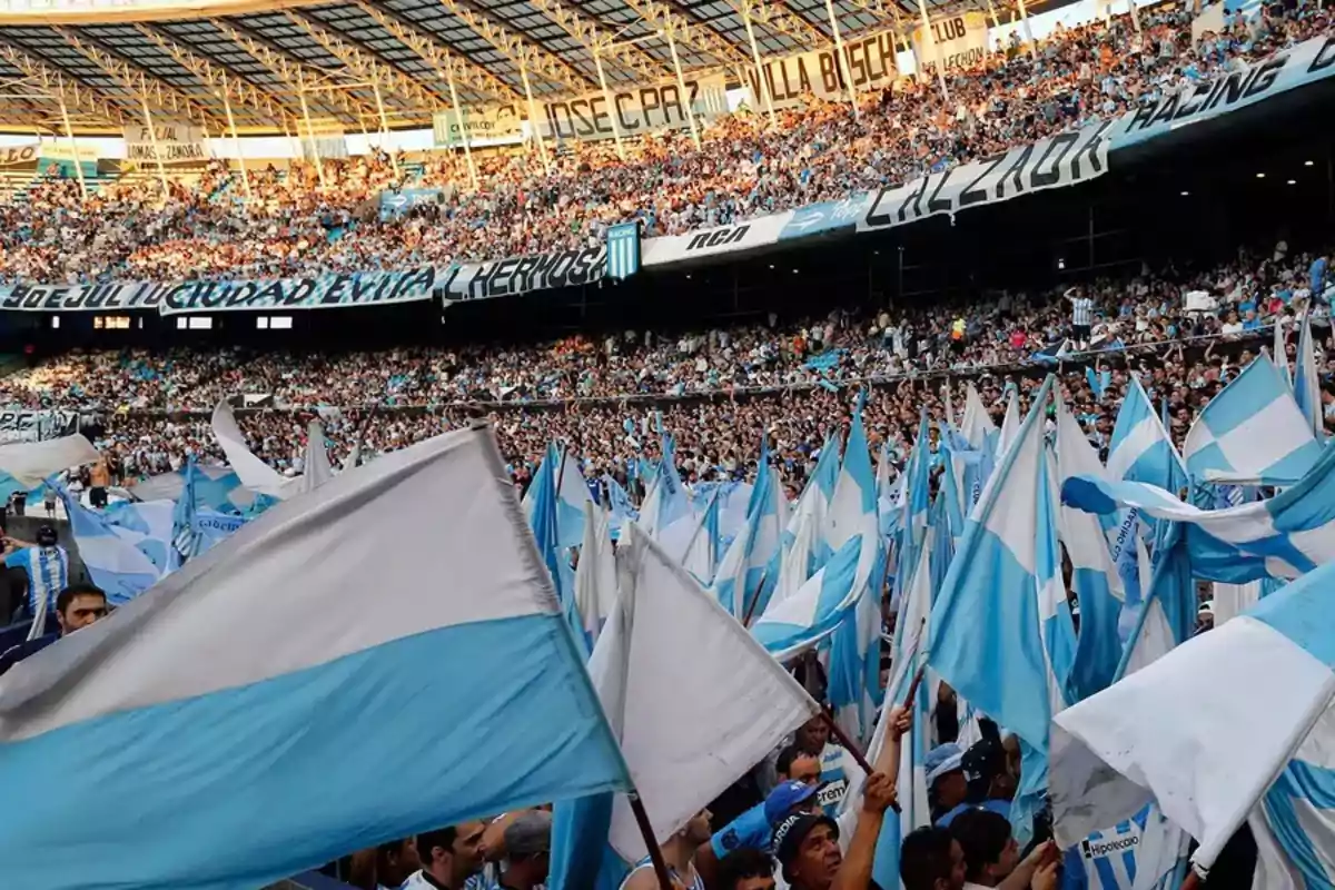 Aficionados en un estadio de fútbol ondean banderas celestes y blancas mientras las gradas están llenas de espectadores.