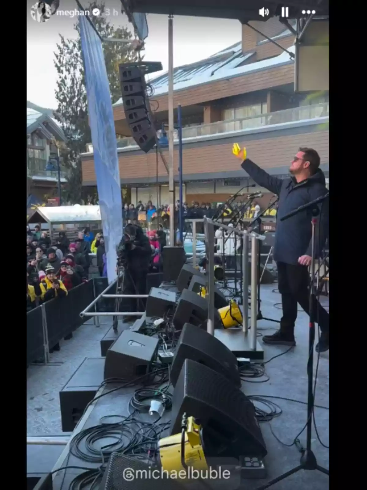 Un hombre en un escenario al aire libre saluda a una multitud mientras sostiene un vaso amarillo, rodeado de equipo de sonido y con un edificio de fondo.