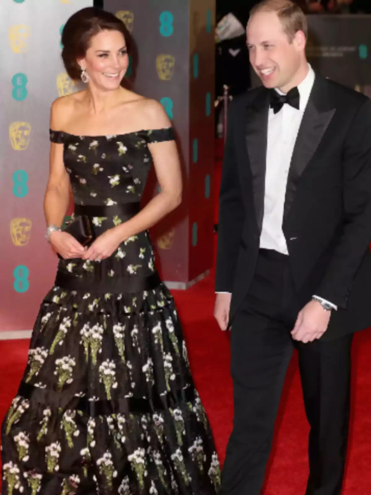 Una pareja elegantemente vestida camina por una alfombra roja, ella lleva un vestido negro con estampado floral y él un esmoquin negro.