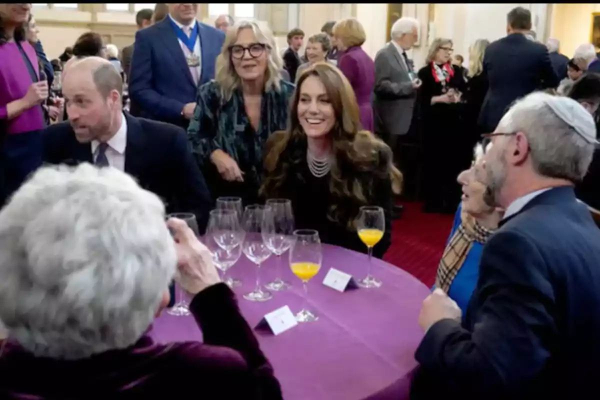 Un grupo de personas conversando y sonriendo alrededor de una mesa cubierta con un mantel morado, con copas de vino y vasos de jugo de naranja.