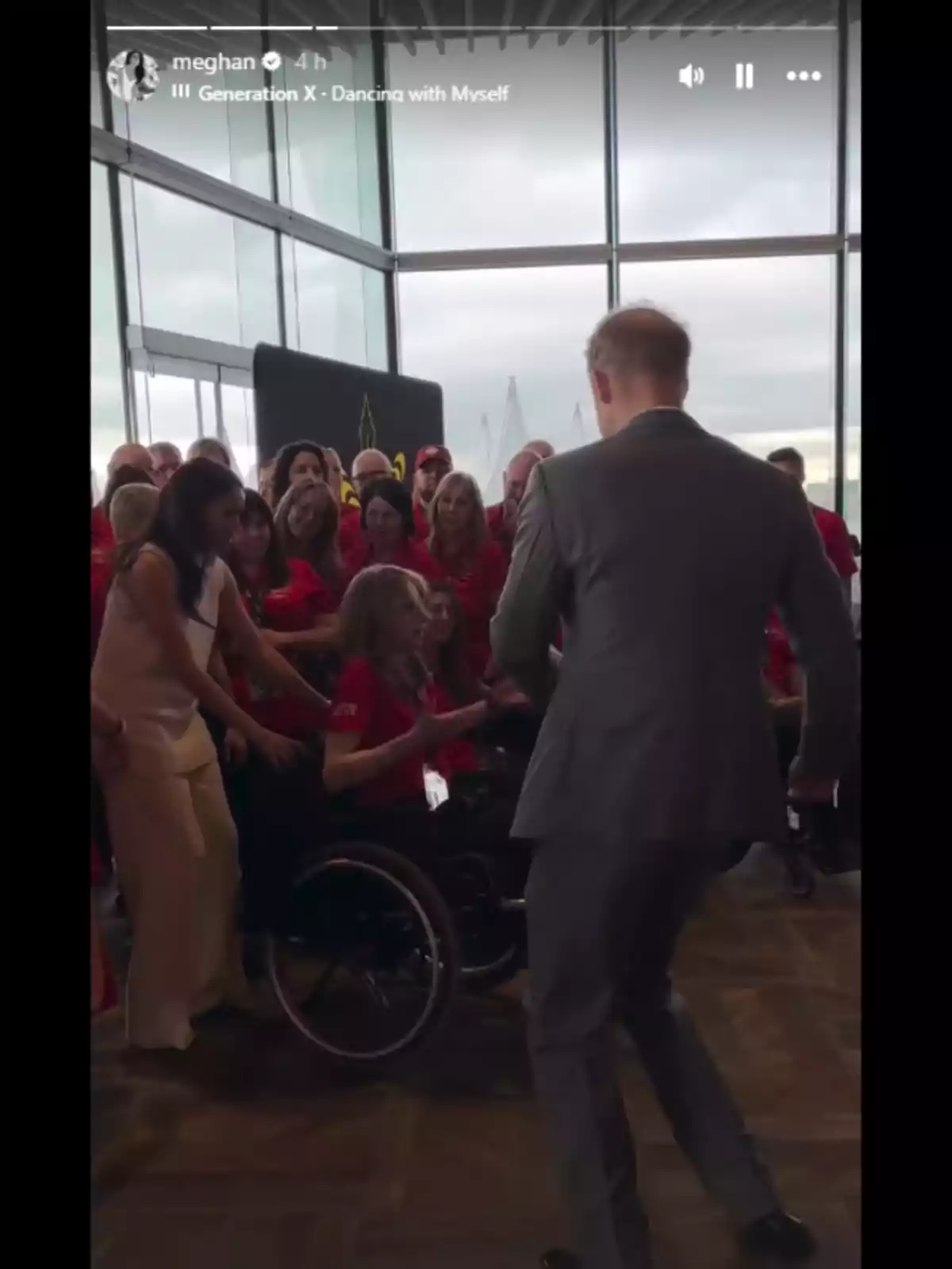 Un grupo de personas se encuentra en una sala iluminada por grandes ventanales, una persona en silla de ruedas está rodeada por otras personas, algunas de las cuales llevan camisetas rojas, mientras una persona de espaldas parece estar interactuando con el grupo.