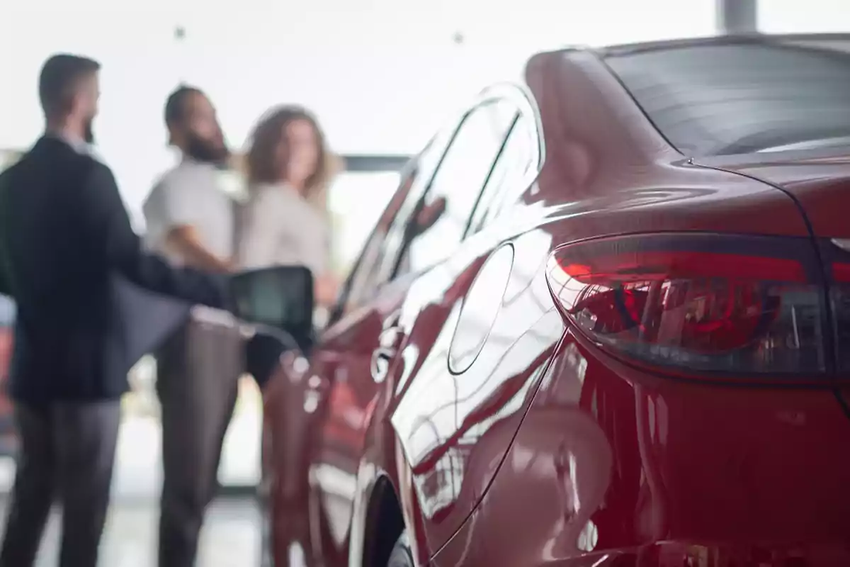 Un coche rojo en un concesionario con tres personas conversando al fondo.