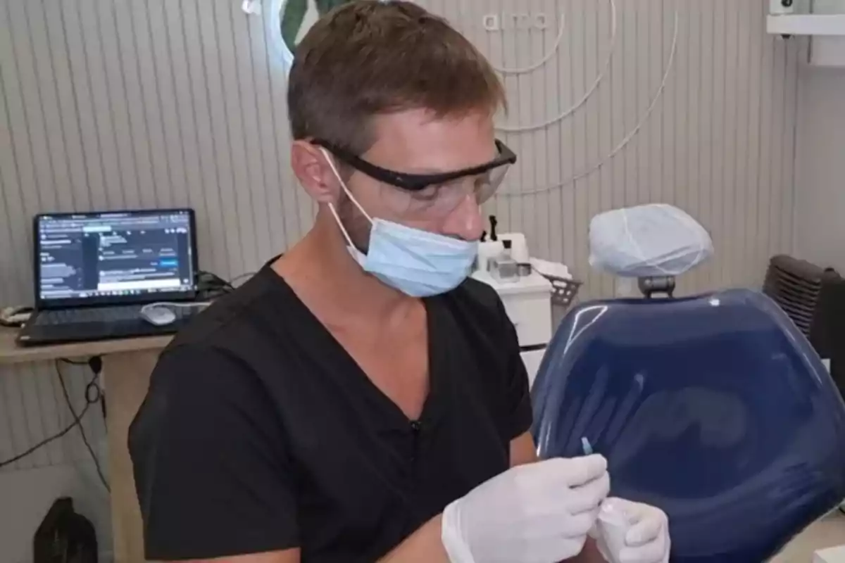 Un dentista con mascarilla y gafas protectoras trabaja en un consultorio dental con una silla azul y una computadora portátil en el fondo.