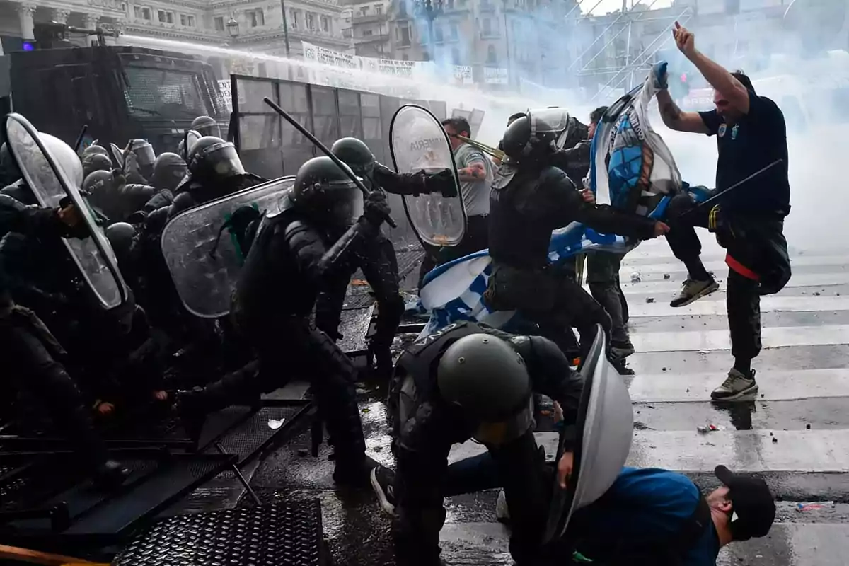 En la imagen se observa un enfrentamiento entre policías antidisturbios y manifestantes en una calle urbana con humo en el ambiente.