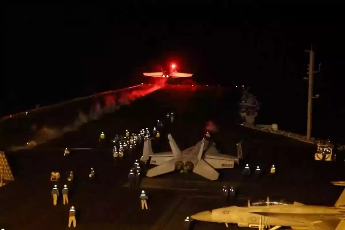 Un avión despegando de un portaaviones en la noche con personal en la cubierta iluminada.