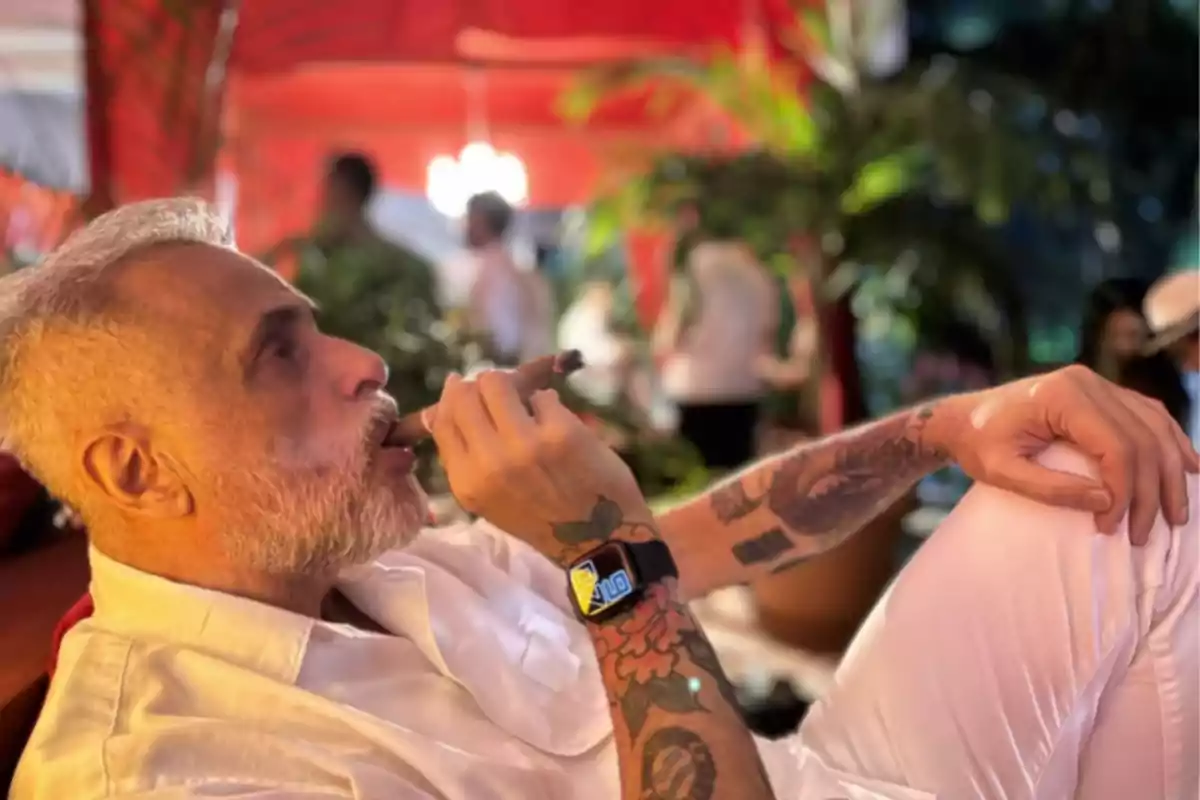 Hombre con barba y tatuajes disfrutando de un cigarro en un ambiente relajado y colorido.