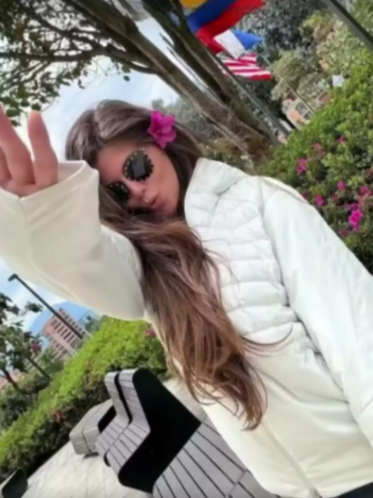 A person with sunglasses and a flower in their hair poses outdoors with flags and greenery in the background.