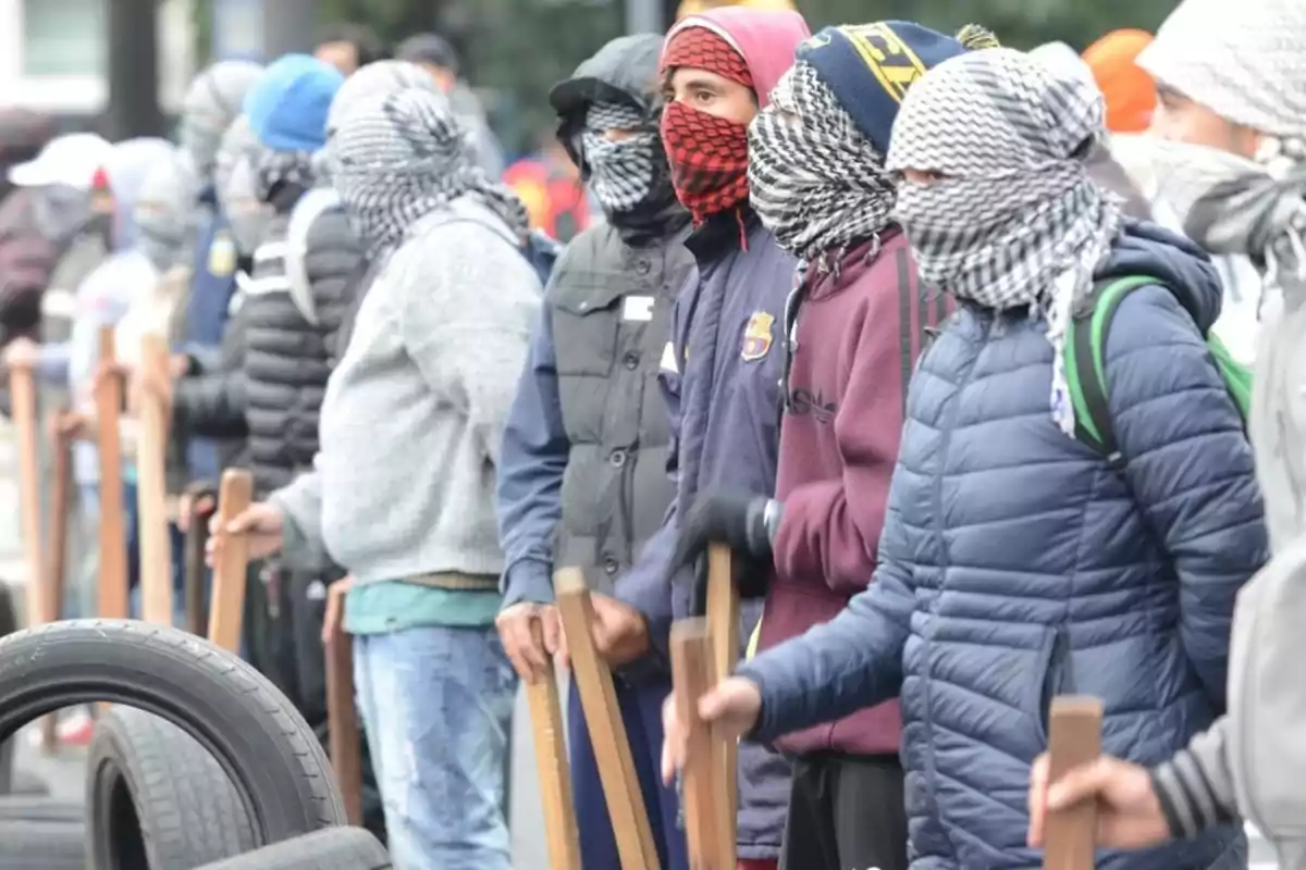 Un grupo de personas con el rostro cubierto sosteniendo palos en una fila.