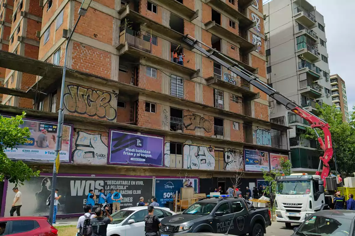 Un edificio en construcción con grafitis en la fachada y un camión con grúa extendida mientras varias personas y policías están en la calle.