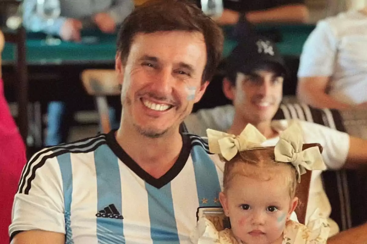 Un hombre sonriente con una camiseta de fútbol de Argentina sostiene a una niña pequeña con la cara pintada y un lazo en el cabello.