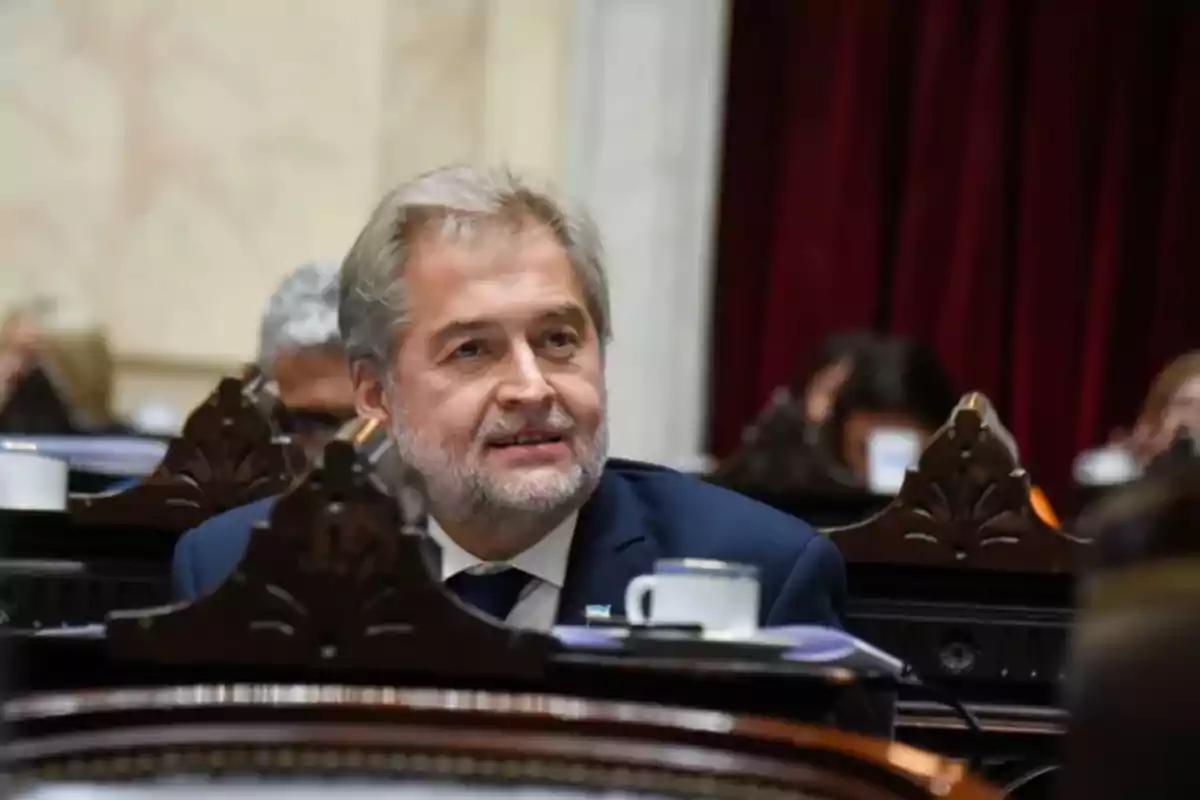 Un hombre de cabello canoso y barba sentado en un entorno formal con cortinas rojas al fondo.