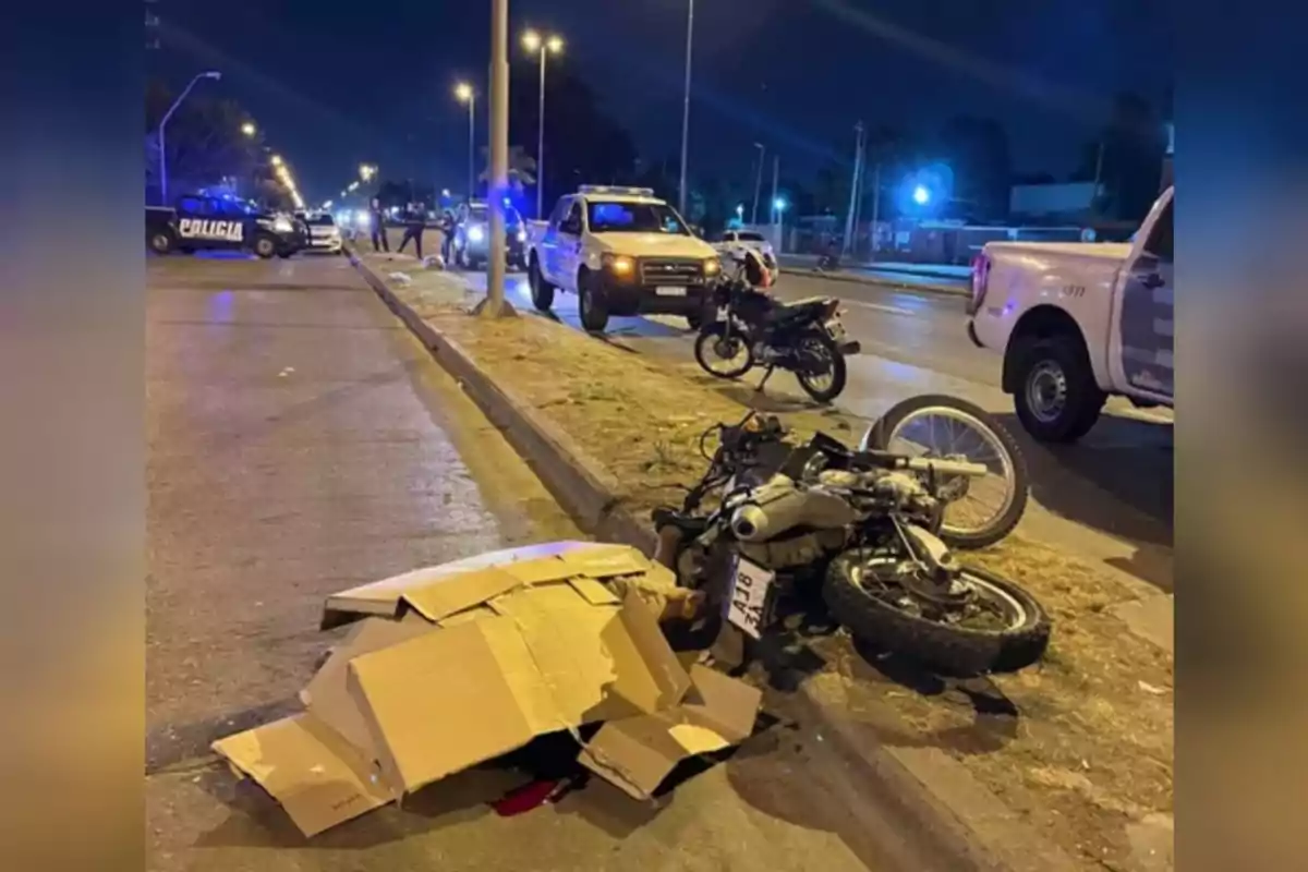 Una escena nocturna muestra un accidente de motocicleta en una avenida con presencia de vehículos policiales y una moto caída en el suelo.