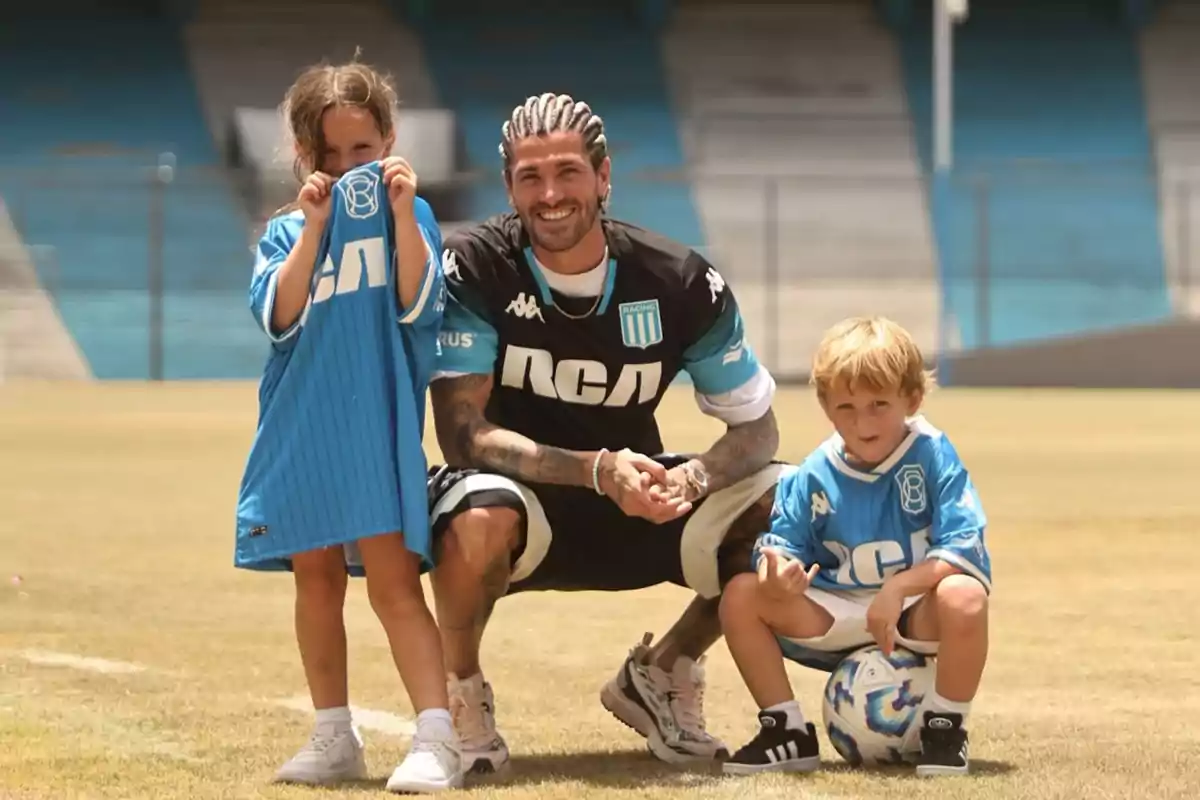 Un hombre sonriente con trenzas posa en un campo de fútbol junto a dos niños que visten camisetas de un equipo deportivo.