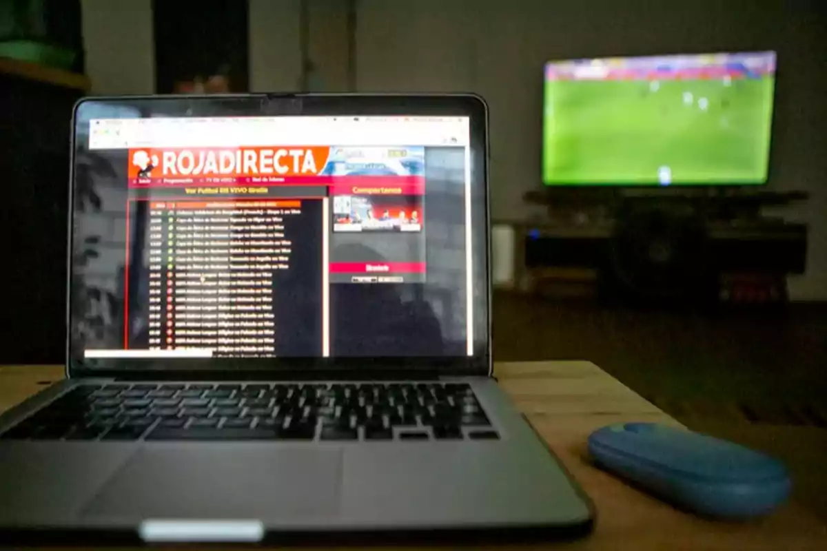 Una laptop muestra un sitio web de transmisión de deportes mientras un televisor en el fondo transmite un partido de fútbol.