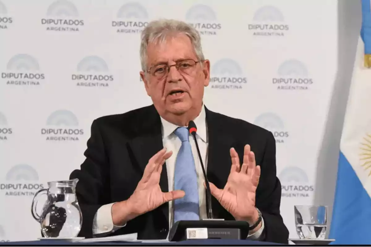 Un hombre mayor con traje y corbata azul hablando en una conferencia de prensa frente a un micrófono con el logo de "Diputados Argentina" de fondo.