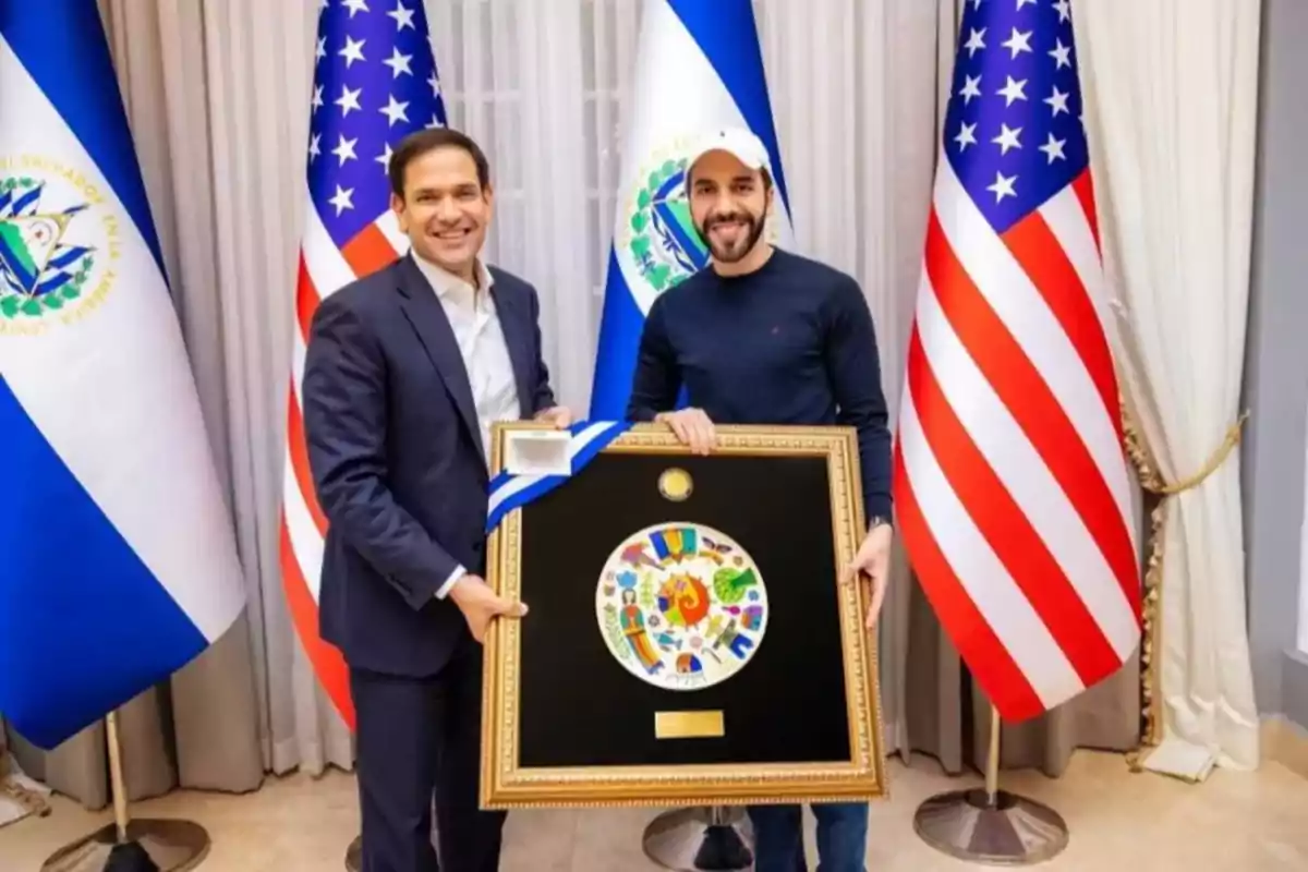 Dos personas posan sonriendo mientras sostienen un cuadro enmarcado, con banderas de El Salvador y Estados Unidos de fondo.