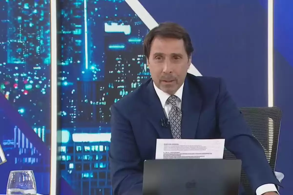 Un hombre con traje azul y corbata está sentado en un estudio de televisión con un fondo de luces de ciudad sosteniendo papeles y frente a una computadora portátil.