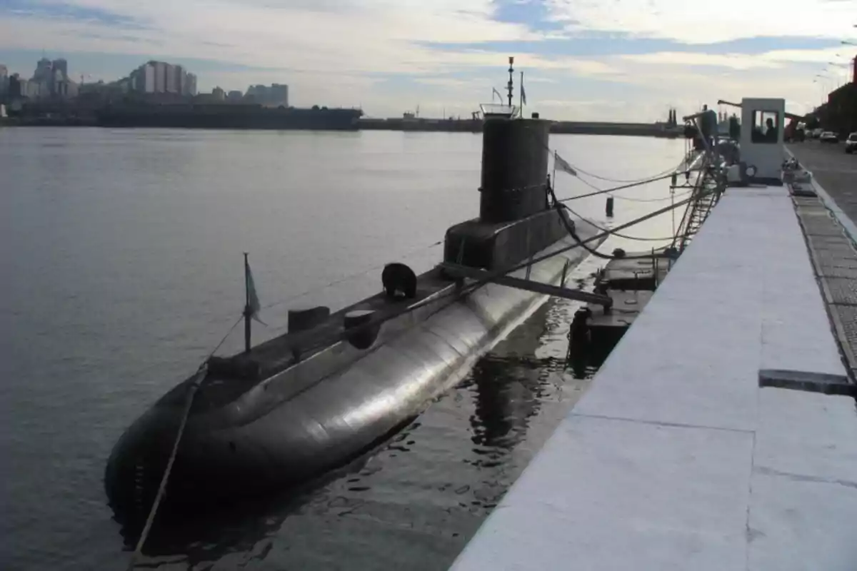 Un submarino atracado en un muelle con edificios al fondo y un cielo parcialmente nublado.