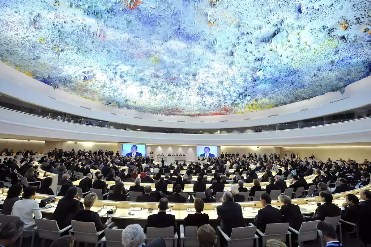 Una gran sala de conferencias con un techo colorido y decorado, llena de personas sentadas en mesas dispuestas en forma circular, con pantallas mostrando a un orador.